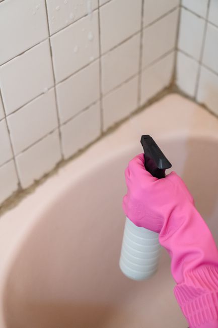 A close-up view of a hand wearing a bright pink rubber glove holding a white spray bottle with a black nozzle, aimed at the grout and ceramic tile corner of a bathroom bathtub. The white tiles on the 